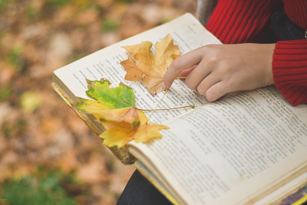 Hand pointing at an open book with autumn leaves scattered over the pages.